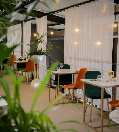 Intérieur de restaurant moderne avec des chaises vertes et orange autour des tables, entouré de plantes et de rideaux blancs vaporeux.