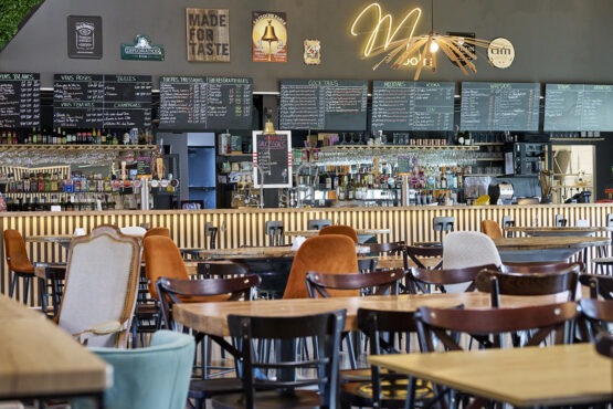 Intérieur chaleureux de café avec des tables en bois et un mélange de sièges. On aperçoit un bar bien approvisionné et des panneaux indiquant les menus