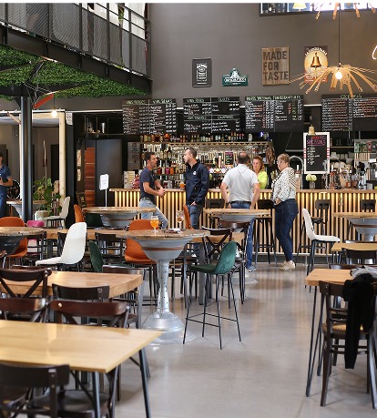 Un café spacieux et moderne, avec des tables et des chaises en bois, où quelques clients sont attablés près du comptoir. Les panneaux indiquant le menu et l'éclairage d'ambiance créent une atmosphère accueillante.