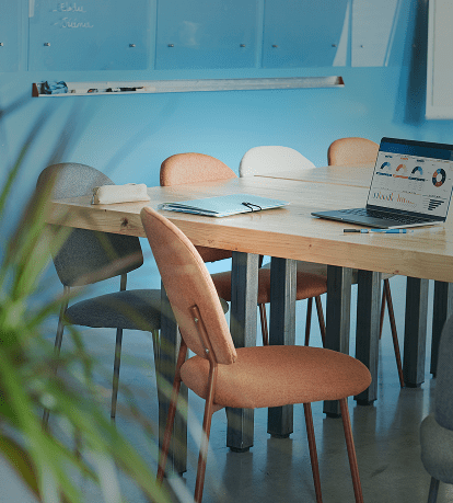 Salle de réunion moderne aux murs bleus, avec une table en bois entourée de chaises de styles variés. Un ordinateur portable ouvert sur la table, une plante à feuilles au premier plan.