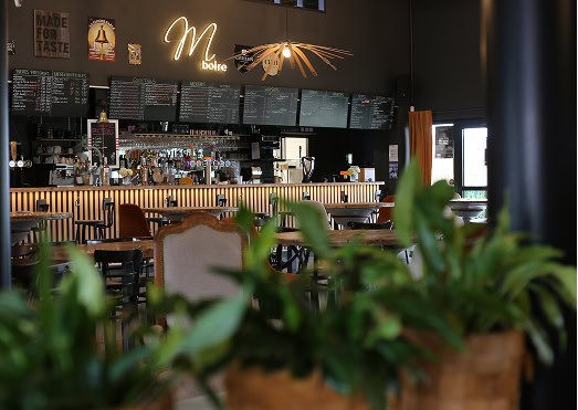 L'intérieur chaleureux du bar, baigné d'une lumière tamisée, est agrémenté d'un comptoir en bois, d'une enseigne lumineuse indiquant « M boire » et de panneaux présentant le menu. Les plantes vertes au premier plan apportent une touche de fraîcheur.